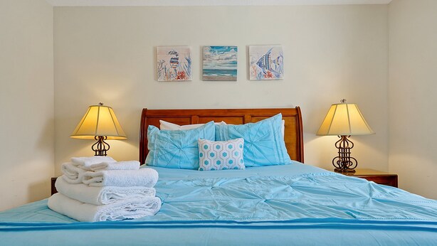 A neatly made bed with blue bedding and white towels, two bedside lamps, and three wall art pieces above the bed. - 7407 Excitement Drive, Kissimmee, Unit A, 1 BED (Kissimmee)