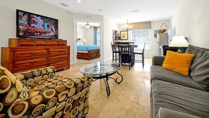 A living room with a TV, a sofa, a glass coffee table, a patterned armchair, and a dining area with chairs and a table. - 7407 Excitement Drive, Kissimmee, Unit A, 1 BED (Kissimmee)