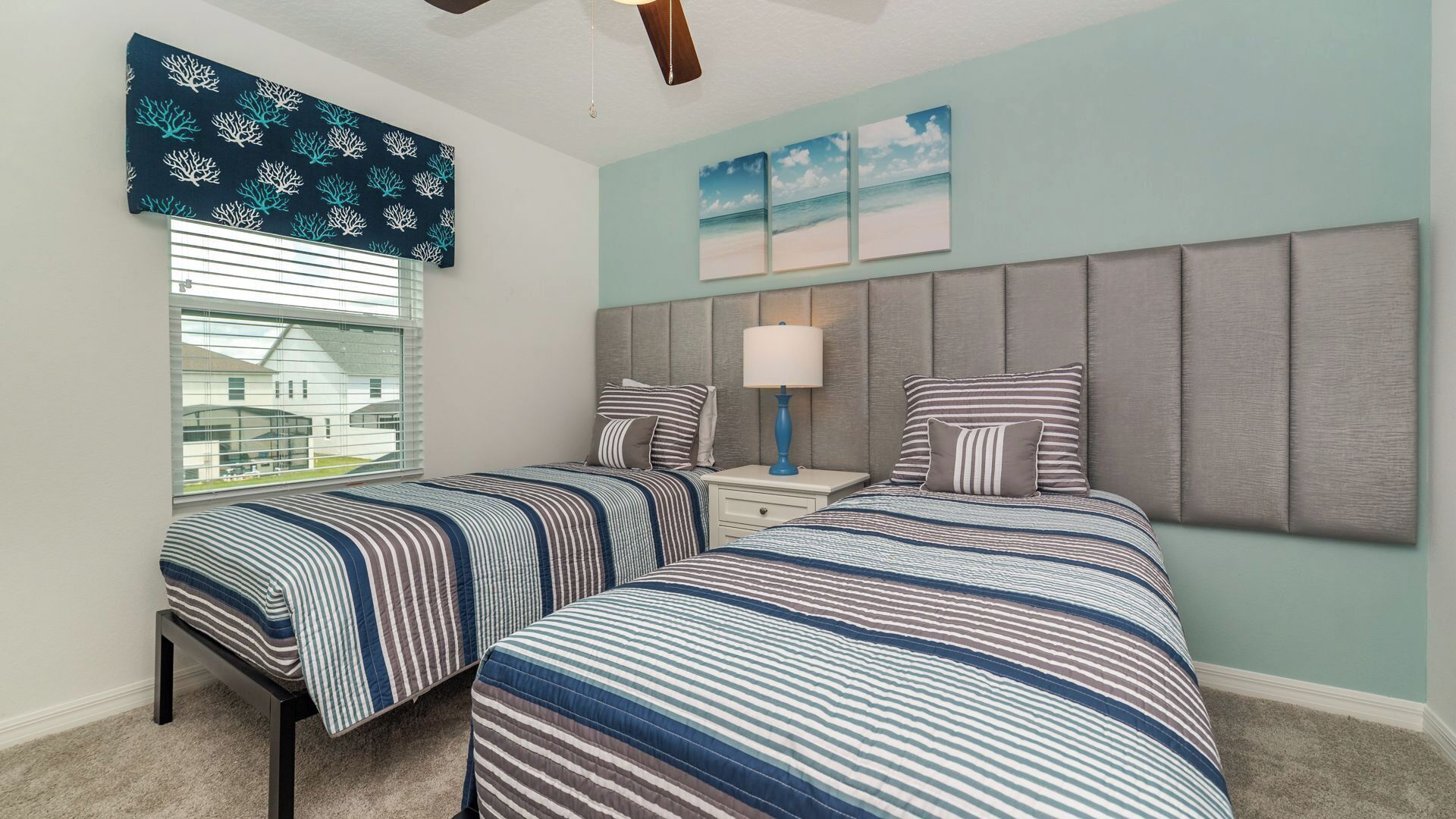 A bedroom with two beds, a window with blinds, a ceiling fan, and a wall with three framed pictures.