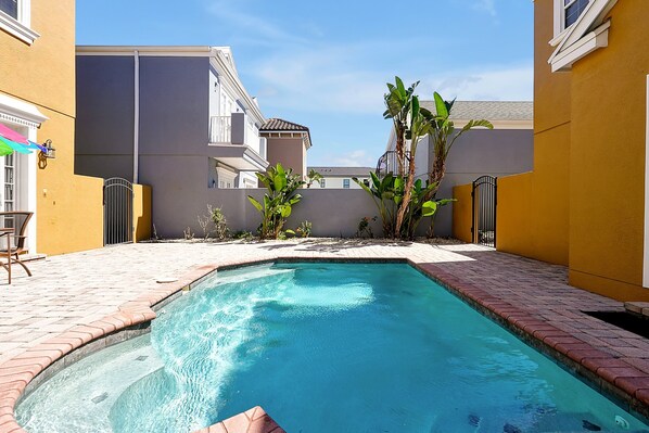 Una piscina con borde de ladrillo, rodeada por un patio con sillas y plantas.
