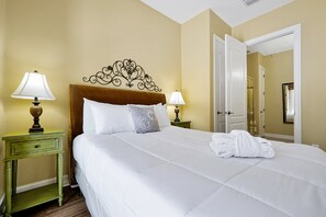 Una cama bien tendida con sábanas blancas y una toalla doblada, dos lámparas de noche, una mesita de noche verde y un cabecero decorativo.