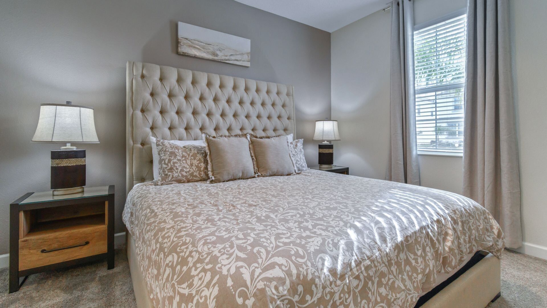 A bedroom with a tufted headboard, a patterned bedspread, two bedside tables with lamps, and a window with blinds.
