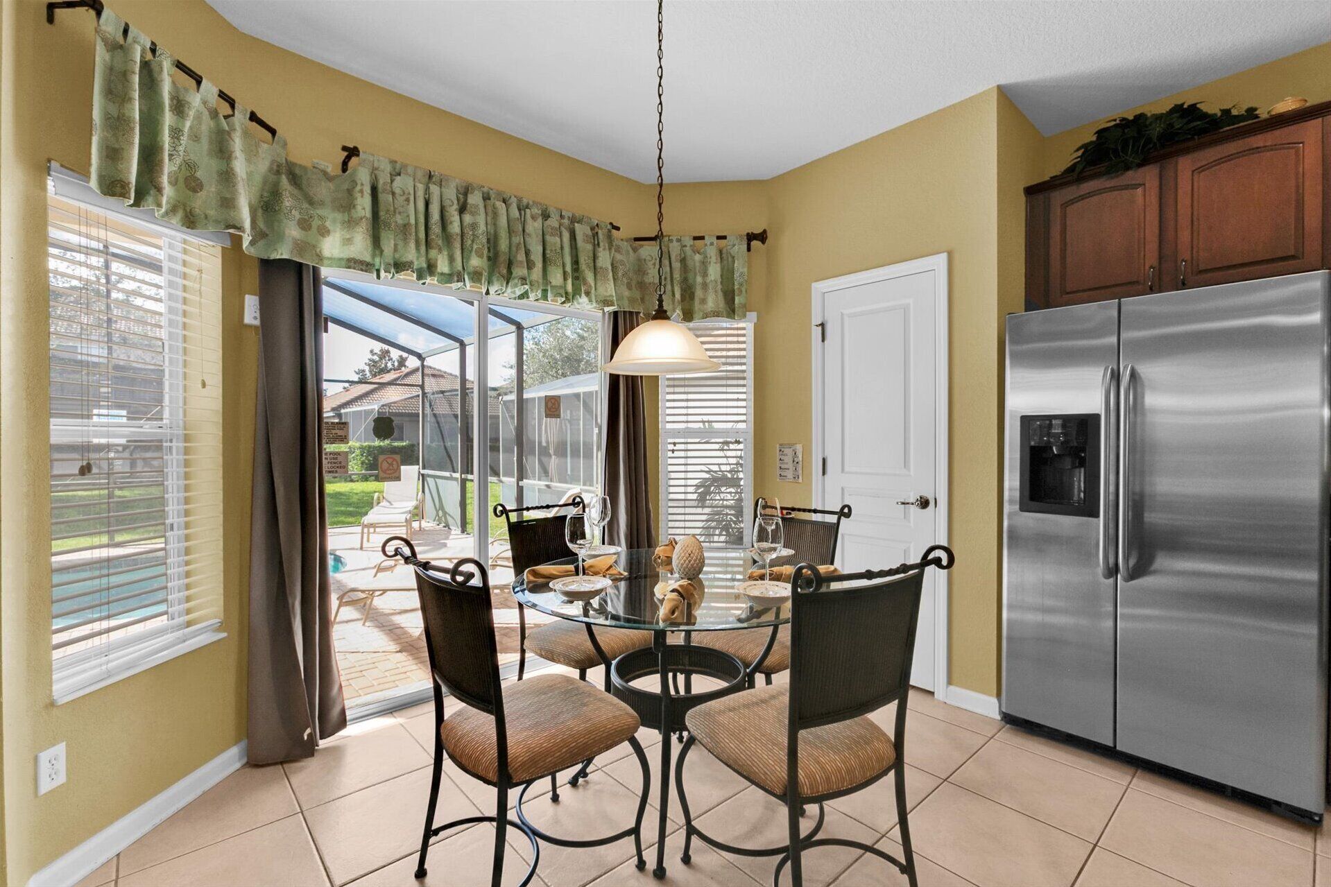 A kitchen with a stainless steel refrigerator, a glass dining table with four chairs, and a view of a backyard through large windows.