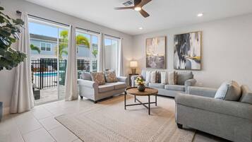 A spacious living room with two sofas, a coffee table, and a ceiling fan. Large windows offer a view of a pool and a building.