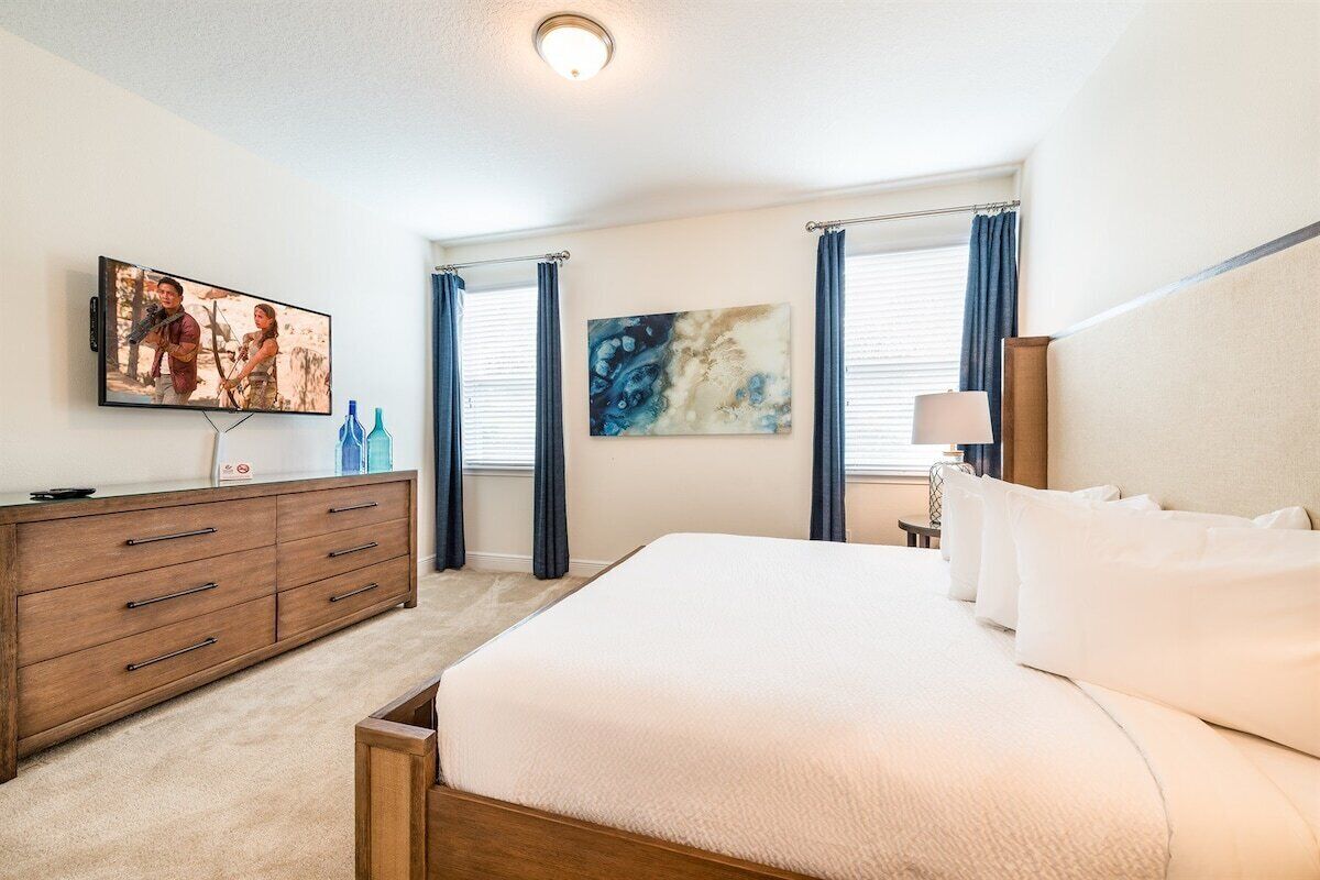 A hotel room with a large bed, a wooden dresser, a flat-screen TV, and two windows with curtains.