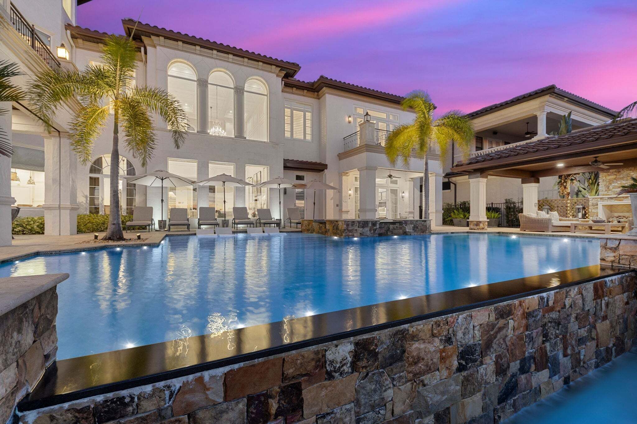 A swimming pool with a stone border, a covered patio area, and a large building with multiple windows and balconies.