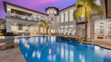 Una casa grande de varios pisos con piscina, mobiliario exterior y palmeras.