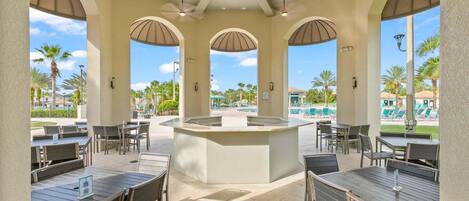 A covered outdoor dining area with tables and chairs, a central island, and views of a pool and palm trees.