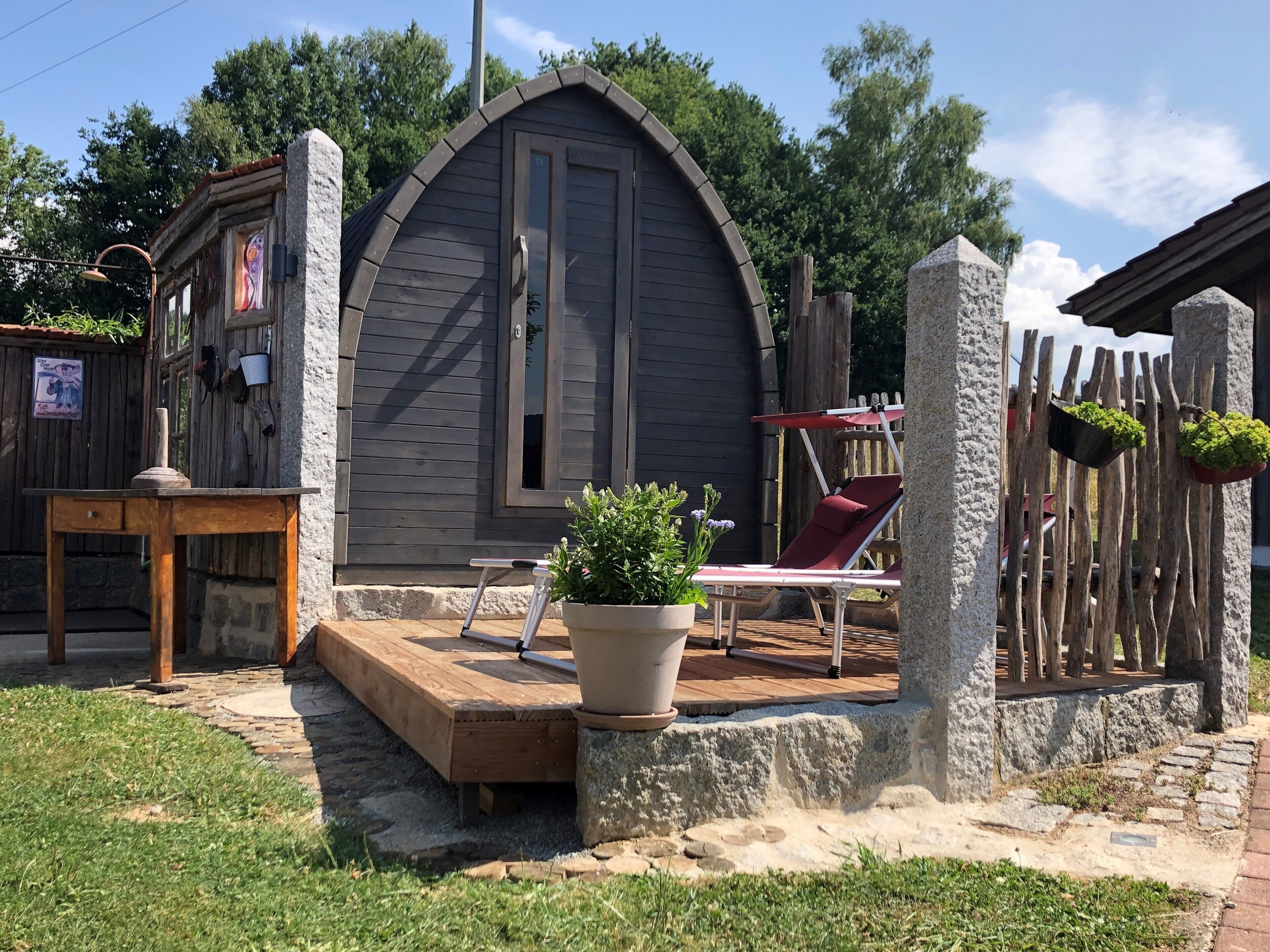 Une cabane en bois rustique, dotée d’un toit en pente, d’un petit porche avec une table et des chaises, et d’une plante en pot.