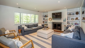 A modern living room with a grey sofa set, a white ottoman, and a fireplace. There are bookshelves with decorative items and a flat-screen TV mounted above the fireplace. - Cozy 6-bedroom beach house with pool, hot tub & AC in the Hamptons (Quogue)