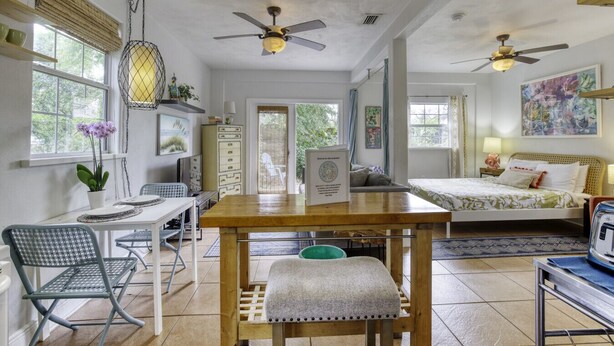 A spacious room with a wooden dining table, two ceiling fans, a bed with a green bedspread, and a large window with a view of greenery. - 1 Bedroom Spanish Bungalow Guest House (Holly Hill)