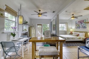 Une pièce spacieuse avec une table à manger en bois, deux ventilateurs de plafond, un lit recouvert d’une couverture verte et une grande fenêtre donnant sur un paysage verdoyant.