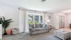 A modern living room with a grey sofa, a white ottoman, and a large window with white curtains. - Seminole Pool House - Mins to the Beach (Seminole)