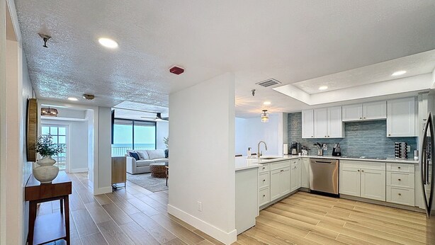 A modern kitchen with white cabinets, a stainless steel refrigerator, and a wooden floor. There is a dining area with a table and chairs visible in the background. - 20002 Gulf Blvd, Penthouse 5 (Indian Shores)
