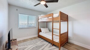 A room with a bunk bed, a ceiling fan, a window with blinds, a television on a stand, and a patterned rug. - 20002 Gulf Blvd, Penthouse 5 (Indian Shores)