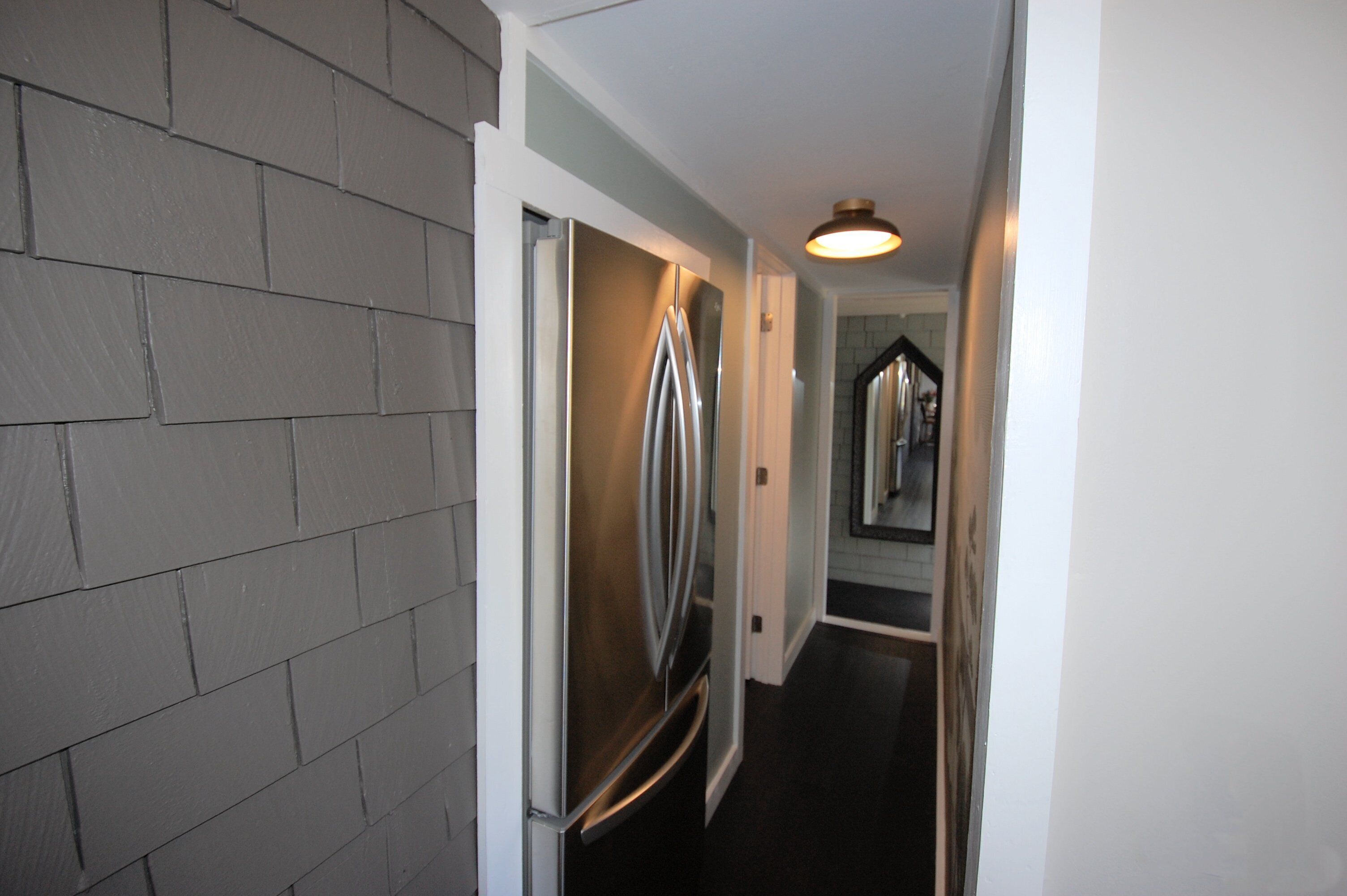 A narrow hallway with a stainless steel refrigerator, a light fixture on the ceiling, and a door leading to another room.