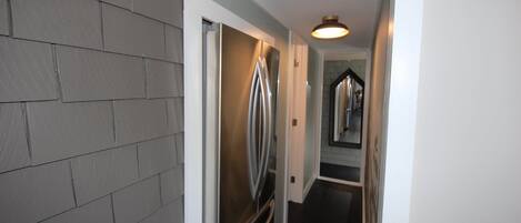A narrow hallway with a stainless steel refrigerator, a light fixture on the ceiling, and a door leading to another room.