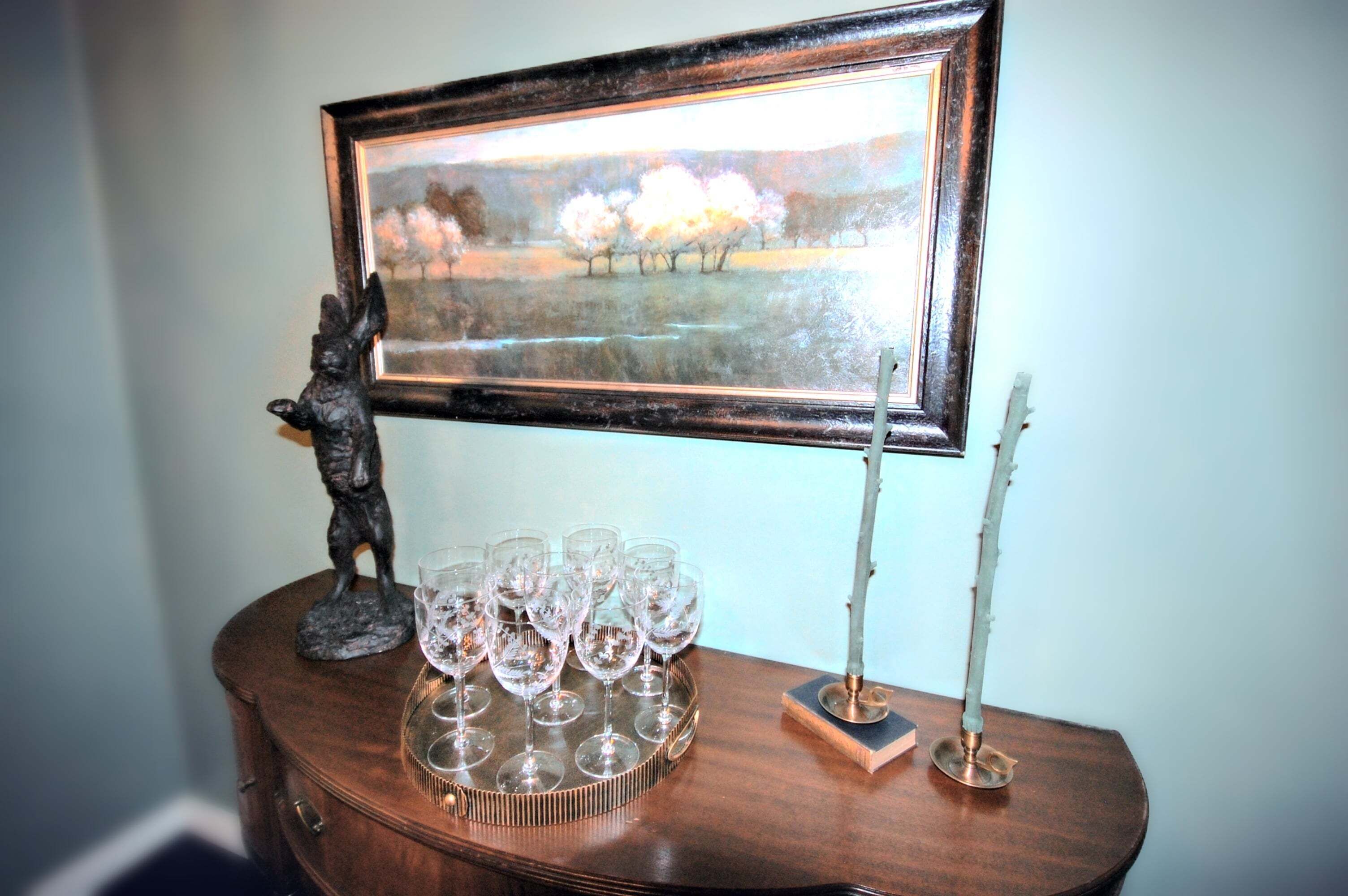 A framed painting, a statue, a tray with wine glasses, and a candle holder on a wooden table.