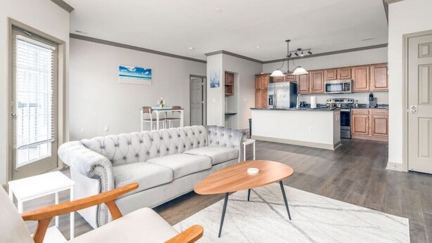 A modern living room with a grey sofa, wooden coffee table, and a dining area with a small table and chairs. A kitchen with wooden cabinets and stainless steel appliances is visible in the background. - Affordable 1-bedroom Apartment Keystone (Indianapolis)