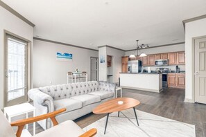 Un salon moderne avec un canapé gris, une table basse en bois et un coin repas comprenant une petite table et des chaises. On aperçoit à l’arrière-plan une cuisine avec des meubles en bois et des appareils électroménagers en acier inoxydable.