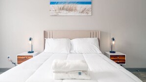 A neatly made bed with white linens, two bedside tables with lamps, and a beach-themed painting on the wall. - Affordable 1-bedroom Apartment Keystone (Indianapolis)