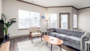 A modern living room with a grey sofa, a wooden coffee table, and a white armchair. There are large windows with blinds, a floor lamp, and a potted plant. - Affordable 1-bedroom Apartment Keystone (Indianapolis)