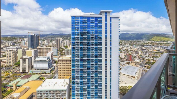 A high-rise building with a modern glass facade, surrounded by a cityscape with various other buildings and a mountainous background. - Studio Ocean View & Balcony 1118 (Honolulu)