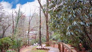 A wooden pathway leading through a forested area with a view of a lake and a wooden bridge. - Highlands Hideout (Highlands)