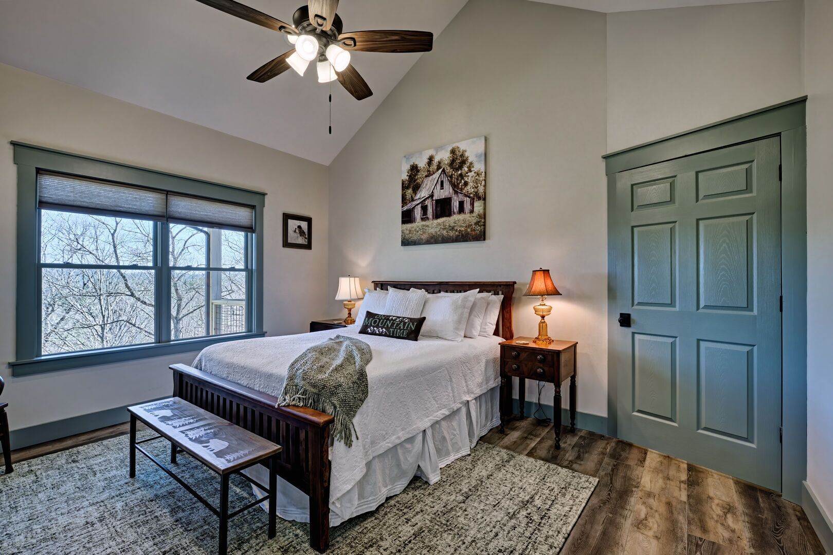 A bedroom with a bed, two bedside tables, a nightstand, a bench, a window, a door, and a ceiling fan.