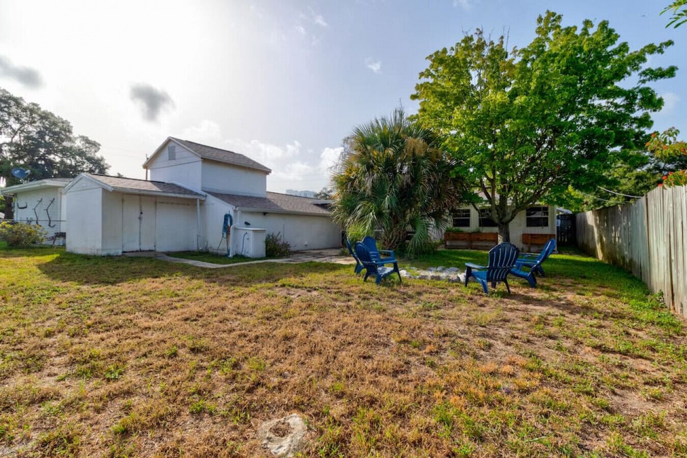 A backyard with a white house, two blue lawn chairs, and a tree.