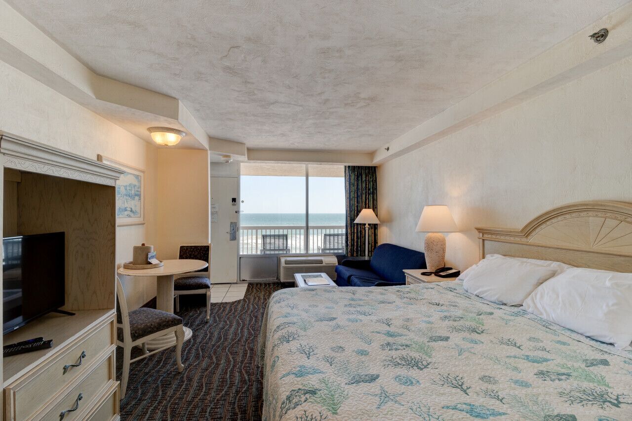 A hotel room with a bed, a TV, a desk, and a balcony with a view of the sea.