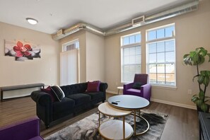 A modern living room with a black sofa, a round coffee table, and a purple armchair. There are large windows with blinds, a radiator, and a painting on the wall. - Private Room Downtown Indy (Indianapolis)