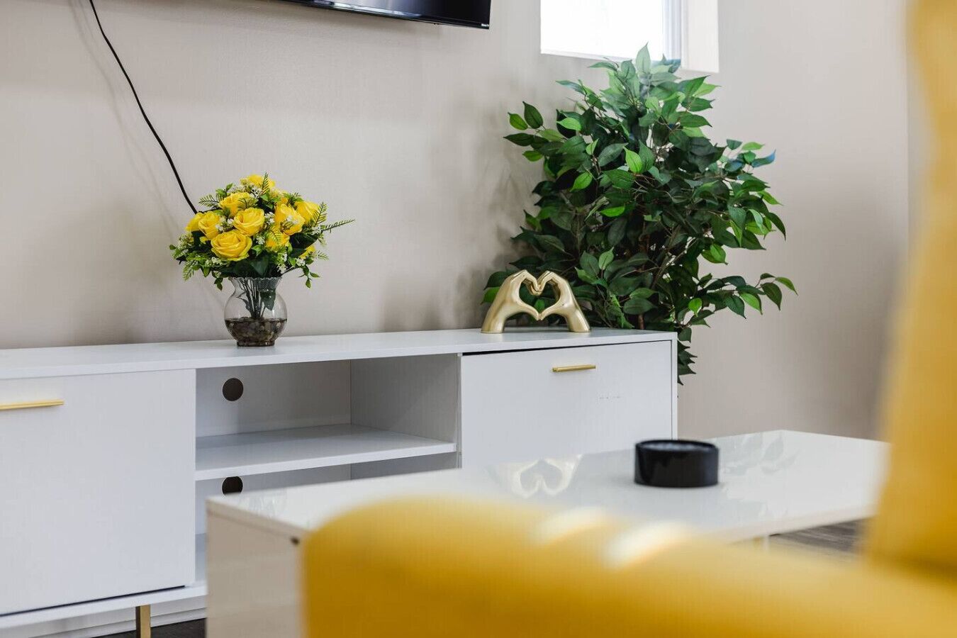 A modern room with a white desk, a vase of yellow flowers, a green plant, and a black object on the desk.