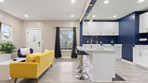 A modern apartment with a yellow sofa, a kitchen with white cabinets, and a large window with curtains. - Modern Comfort Downtown Indy Stylish 3BR Getaway (Indianapolis)