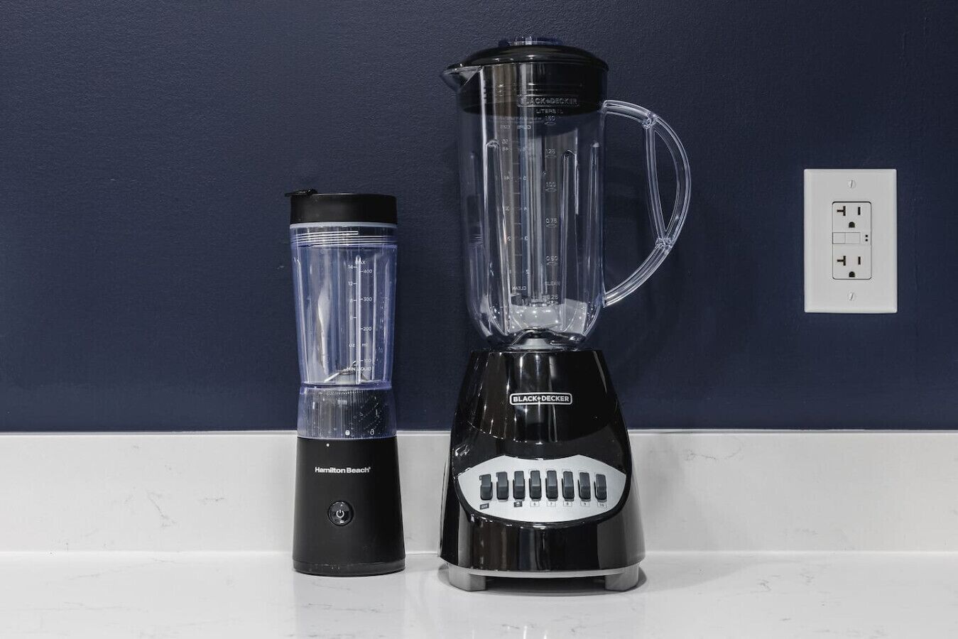 A black Hamilton Beach blender and a black Black+Decker blender on a white countertop against a dark blue wall.