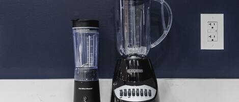 A black Hamilton Beach blender and a black Black+Decker blender on a white countertop against a dark blue wall.
