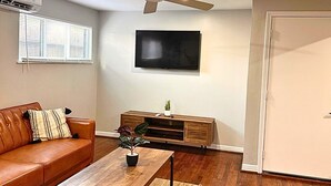A living room with a brown leather sofa, a wooden coffee table, a TV on the wall, a ceiling fan, and a door. - Modern 2 Bed Near Rice University (Houston)
