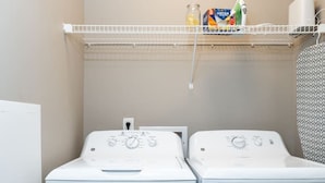 A laundry area with two white front-loading washing machines and a shelf above containing cleaning supplies. - Downtown Indy Private Room (Indianapolis)