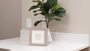 A modern room with a white desk, a framed inspirational quote, a large green plant, and a tufted grey chair. - Downtown Indy Private Room (Indianapolis)