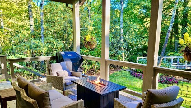A covered outdoor seating area with wicker furniture, a fire pit, and a view of a lush garden. - Dancing Bear Walk to Town (Highlands)