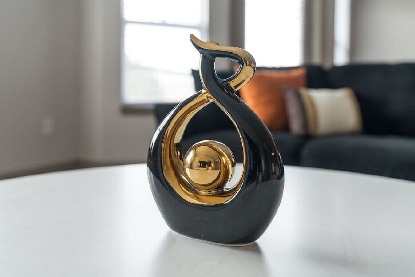 Un objet décoratif noir et doré est posé sur une table blanche, dans une pièce où se trouve un canapé aux couleurs sombres et une fenêtre en arrière-plan.