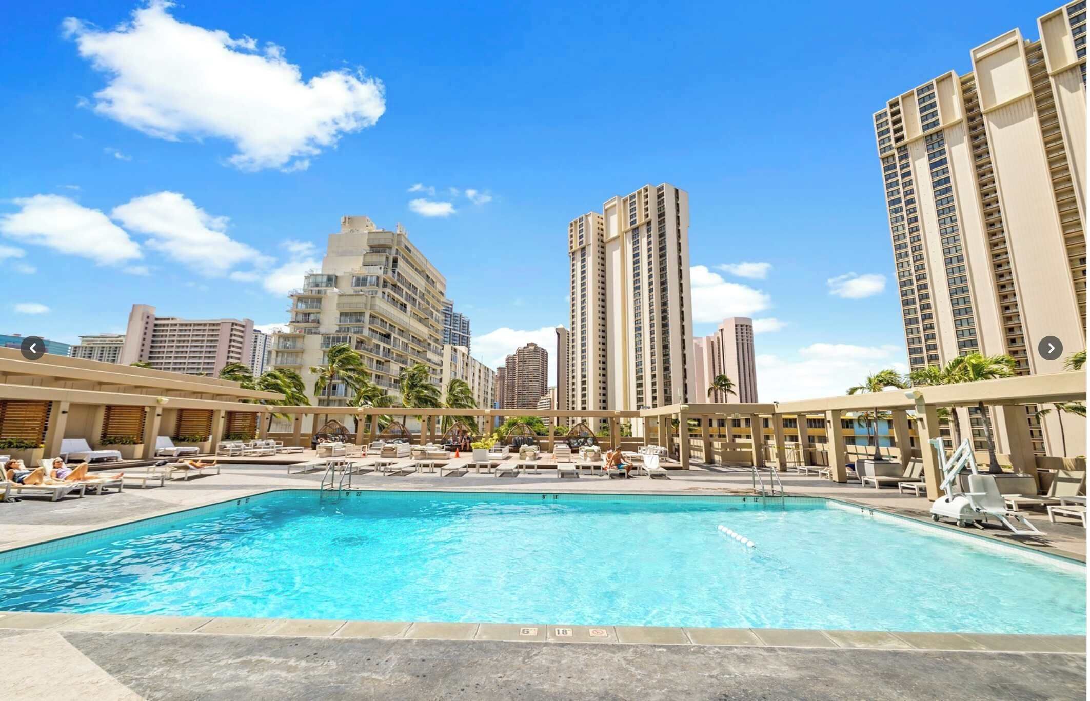 A swimming pool with lounge chairs and umbrellas, surrounded by tall buildings.