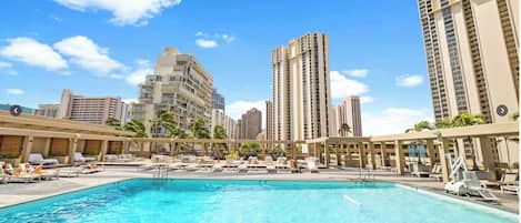 Piscina com espreguiçadeiras e guarda-sóis, rodeada por edifícios altos.