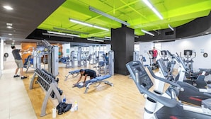 Une salle de sport équipée de divers appareils d’exercice, dotée d’un plancher en bois et de luminaires verts vifs au plafond.