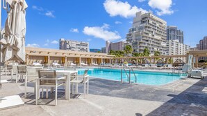 Zona de piscina de hotel con sillones de playa y sombrillas, rodeada de edificios altos.