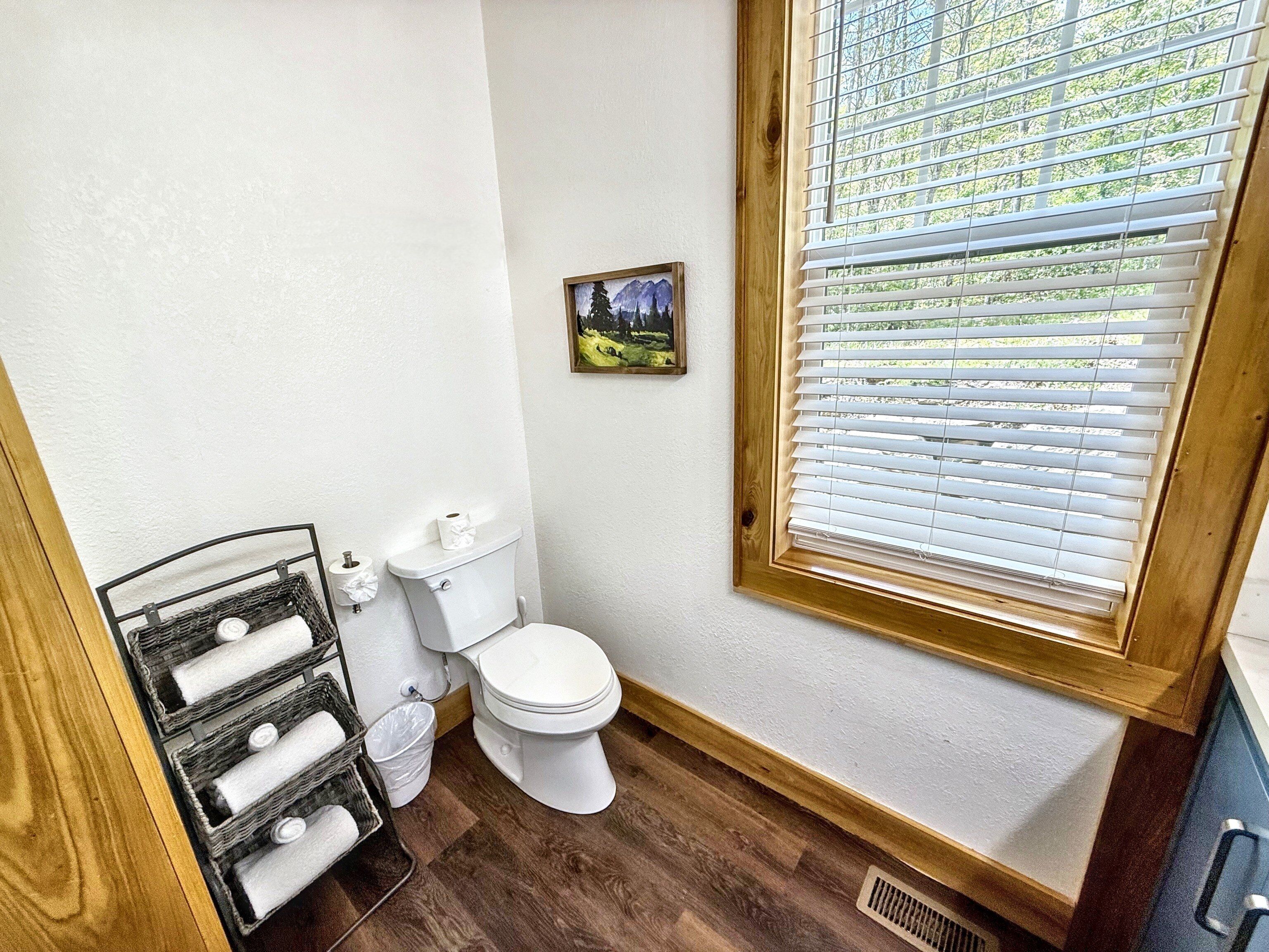 Een kleine badkamer met toilet, een raam met zonwering, een houten vloer en een aan de muur hangend schilderij.