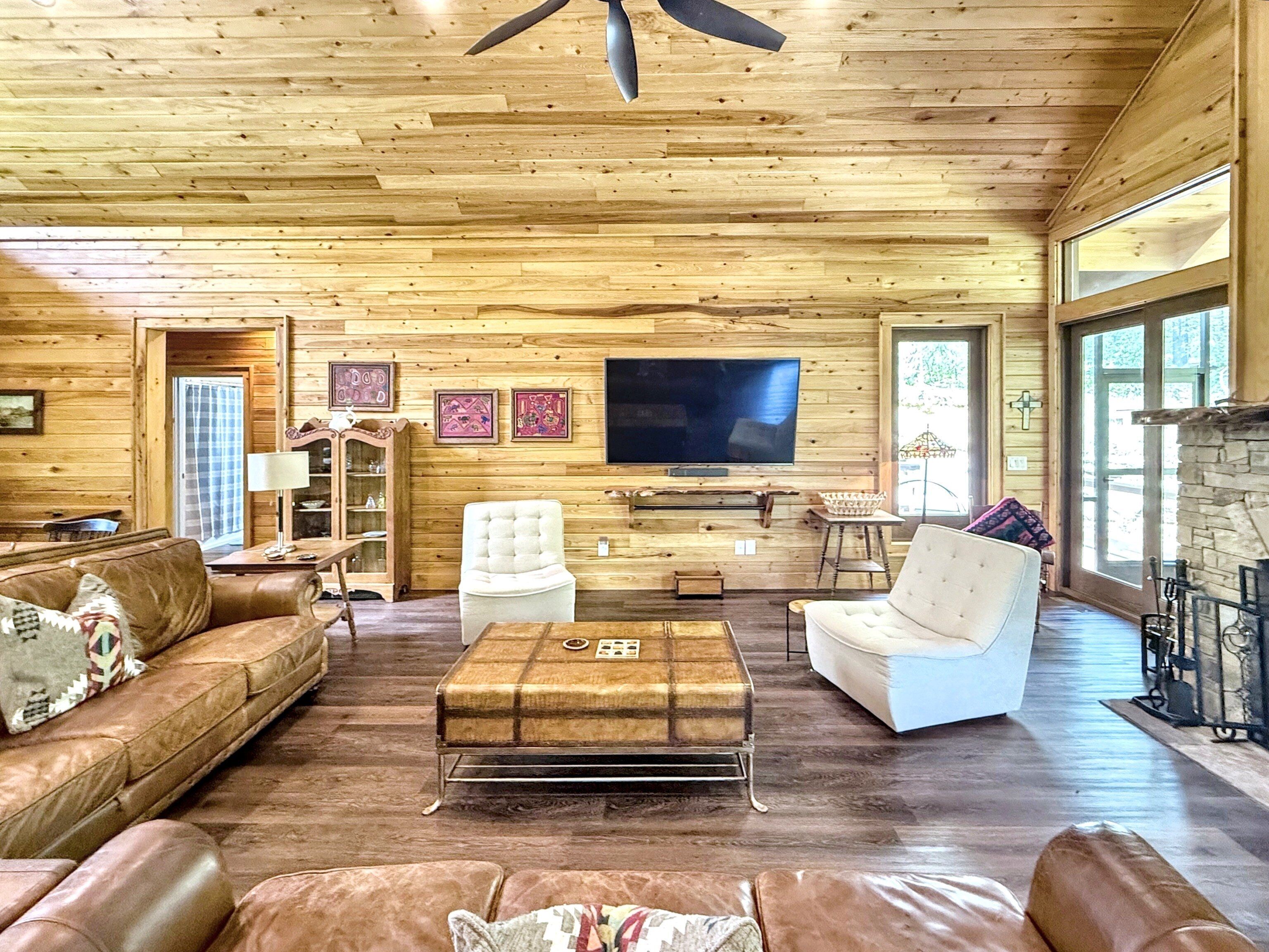 A spacious living room with wooden walls and floors, a large flat-screen TV mounted on the wall, and a fireplace on the right side.