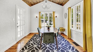 A dining area with a wooden ceiling, a chandelier, a round table with chairs, and a view through the windows. - Spanish Beach Bungalow Mainhouse (Holly Hill)