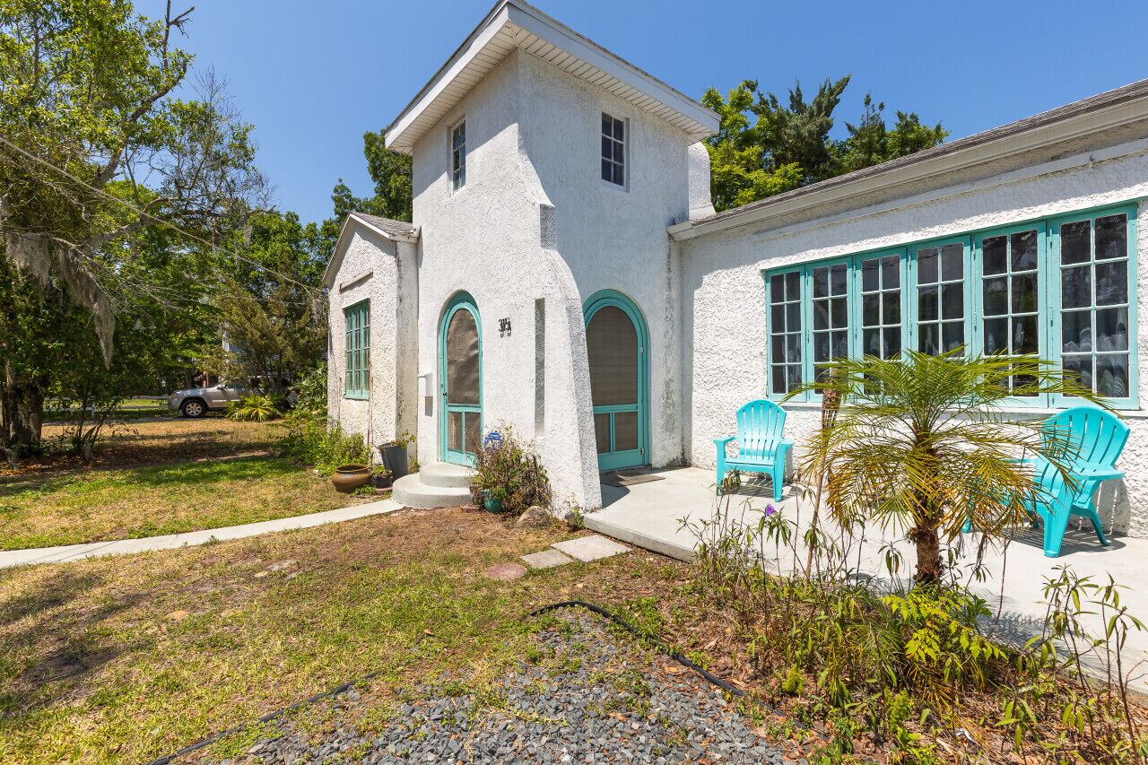 A white building with turquoise accents, a small porch with chairs, and a well-maintained lawn.