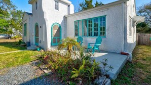 A two-story house with a white exterior and turquoise accents, including windows and doors. There are two outdoor chairs and a small garden area with various plants. - Spanish Beach Bungalow Mainhouse (Holly Hill)